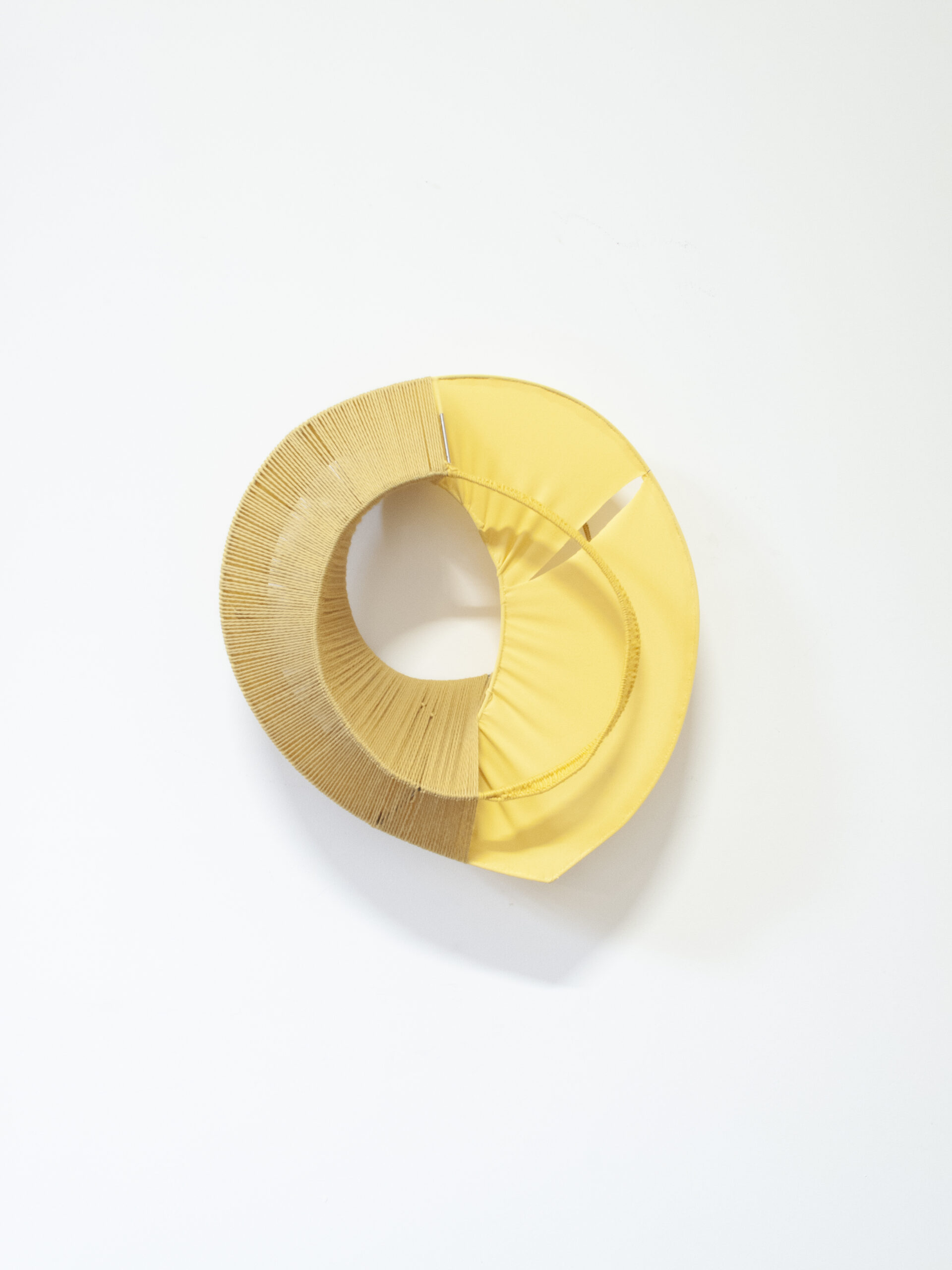 A sculpture installed on a white wall. It is circular in shape. The right half is made of a yellow fabric while the left is made of a straw like material.