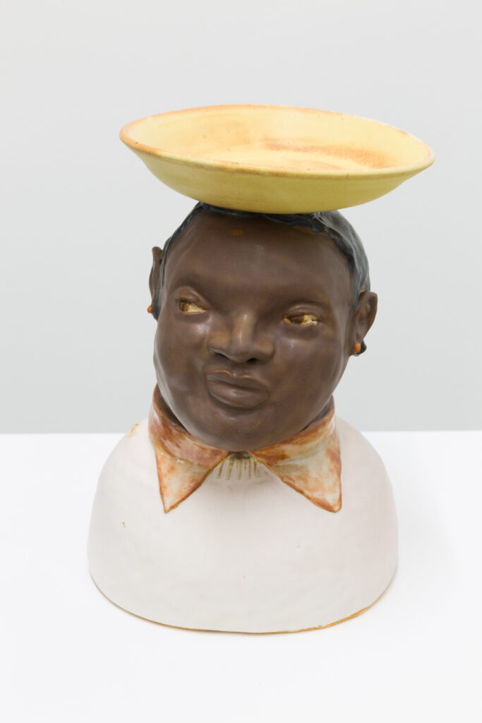 A ceramic sculpture of a person with a yellow bowl on top of their head. The figure is facing the camera, their eyes are looking off to the left.