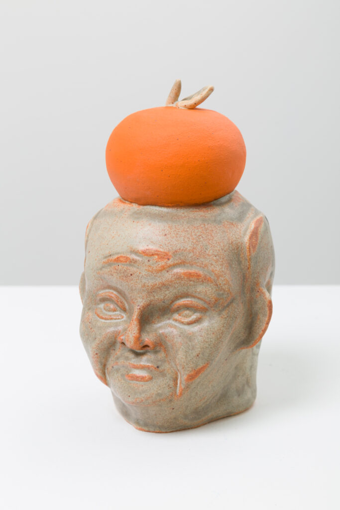 A ceramic bust of a figure with an orange and grey glaze. The figure is angled 3/4 and there is an orange on top of the figure's head