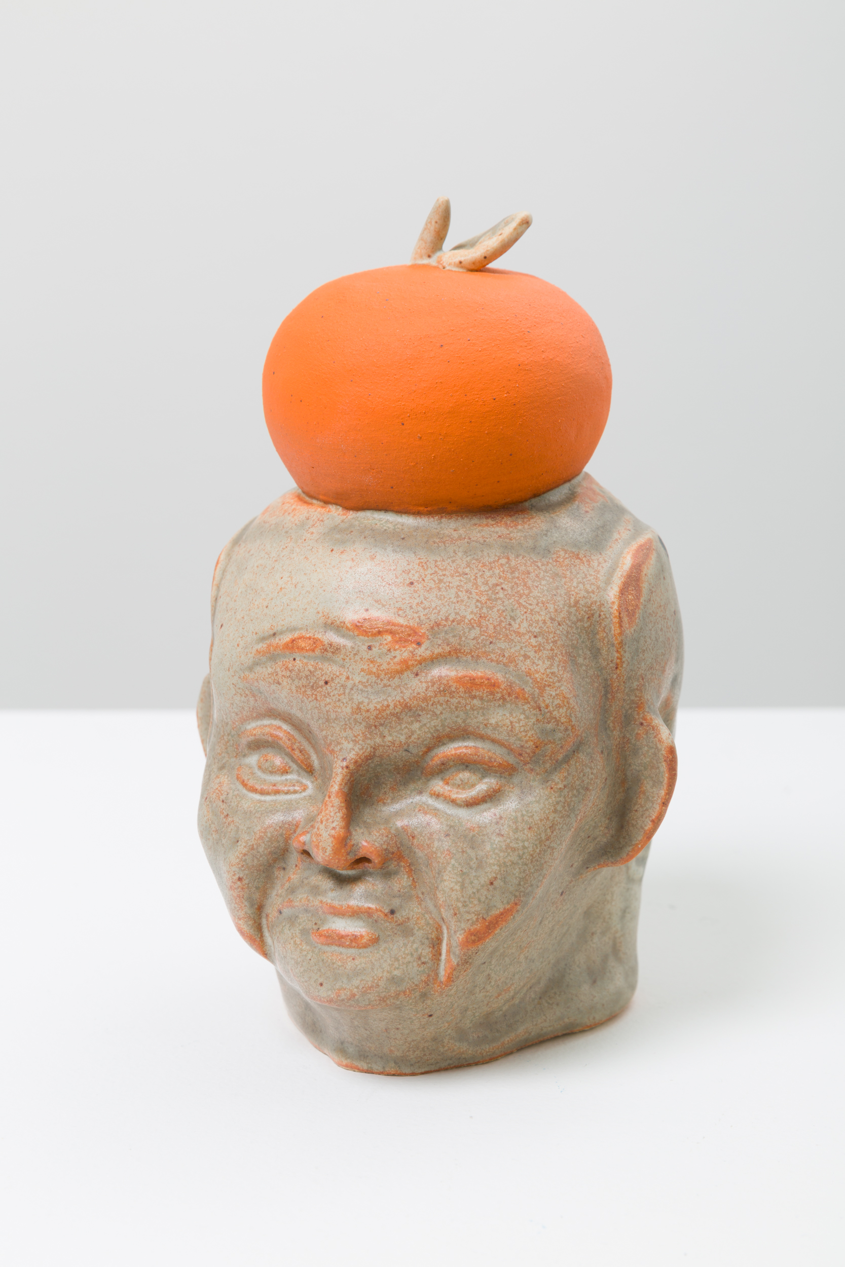 A ceramic bust of a figure with an orange and grey glaze. The figure is angled 3/4 and there is an orange on top of the figure's head