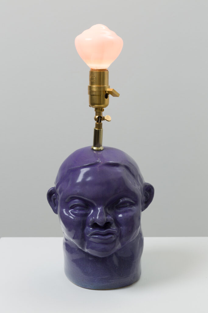 A purple ceramic bust of a figure with a gold light fixtures extending up from the top of the figure's head. The light bulb is glowing light pink and the figure is facing towards from the camera.